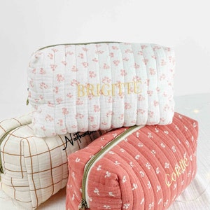 Op de afbeelding: Drie quilted toilettassen met rits. De bovenste tas is wit met roze bloemenprint en de naam "Brigitte" geborduurd in goud. De middelste tas is beige met een bruin ruitpatroon en de naam "Nathalie" geborduurd in zwart. De onderste tas is roze met witte bloemenprint en de naam "Collette" geborduurd in goud.