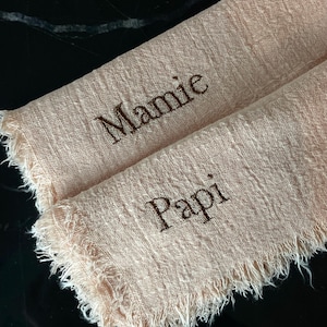Può includere: Due quadrati di tessuto rosa pallido con bordi sfrangiati. Il quadrato superiore ha la parola "Mamie" ricamata in filo marrone. Il quadrato inferiore ha la parola "Papi" ricamata in filo marrone.