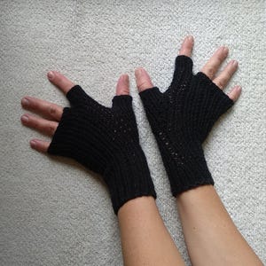 May include: A pair of black fingerless gloves, knitted with a ribbed pattern. The gloves cover the hand up to the knuckles, leaving the fingers free. The gloves are displayed against a light gray background.