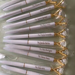 May include: A collection of white pens with gold accents and diamond-shaped tops. Each pen is personalized with a name and title, such as "Pra. Norma Mendes" and "Ob. Marta Soares".