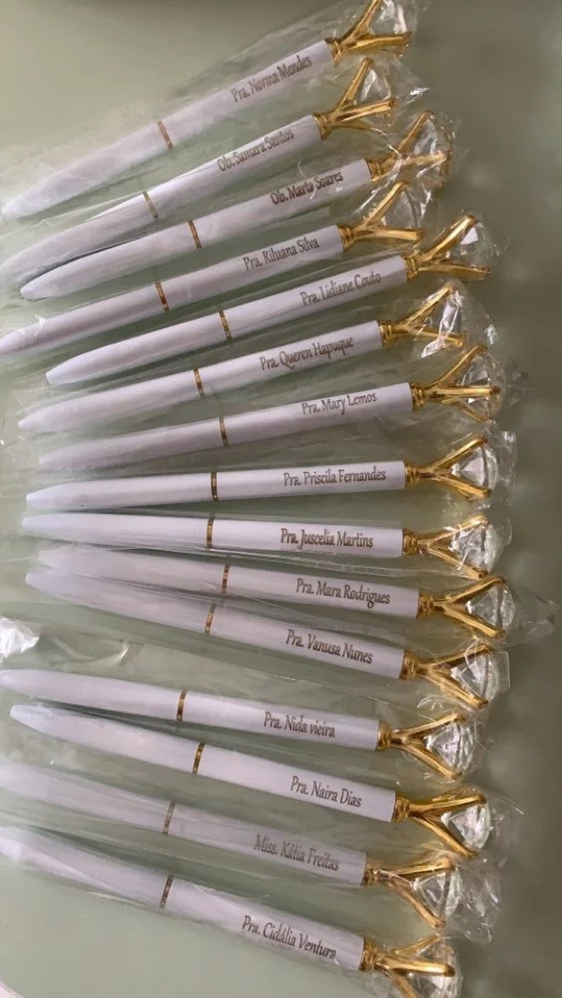 May include: A collection of white pens with gold accents and diamond-shaped tops. Each pen is personalized with a name and title, such as "Pra. Norma Mendes" and "Ob. Marta Soares".