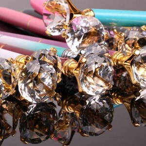 May include: A group of pens with gold-toned tops and large, clear crystal-like embellishments. The pens are arranged in a cluster, with the crystal embellishments facing the camera.