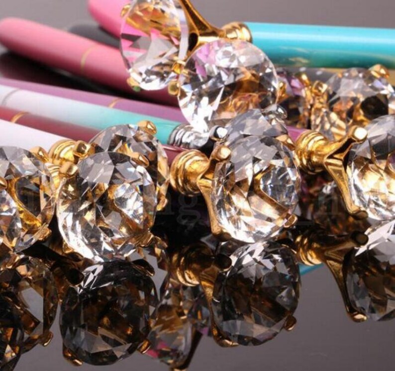 May include: A group of pens with gold-toned tops and large, clear crystal-like embellishments. The pens are arranged in a cluster, with the crystal embellishments facing the camera.