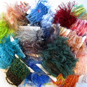 May include: A collection of colorful yarn and fuzzy fibers in various textures, including green, blue, red, and brown. The yarn is arranged in a pile on a white surface.
