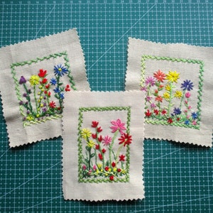 Può includere: Tre quadrati ricamati con diversi motivi floreali. Ogni quadrato presenta una varietà di fiori colorati, tra cui rossi, rosa, gialli e blu, su uno sfondo bianco. I quadrati sono circondati da un bordo verde.