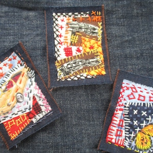 Op de afbeelding: Drie kleine geborduurde pleisters genaaid op een denim achtergrond. De pleisters zijn gemaakt van verschillende stoffen en hebben verschillende ontwerpen, waaronder een auto, een vlinder en een vogel met de tekst "Meet in the World".
