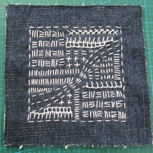 Op de afbeelding: Een donkerblauw denim vierkant met witte stiksels in verschillende patronen, waaronder kruisen, lijnen en zigzaglijnen.