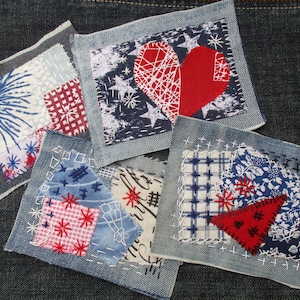 Può includere: Cinque piccoli quadrati di tessuto, ognuno con un diverso design a patchwork, sono cuciti su uno sfondo di denim. I quadrati presentano tessuti rossi, bianchi e blu con vari motivi, tra cui stelle, quadri e disegni floreali.