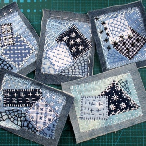 May include: Five small denim squares with different patchwork designs. Each square is made of various blue and white fabrics with different stitching patterns. The squares are arranged on a green cutting mat.