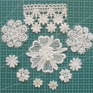 May include: A collection of white lace appliques in various floral designs, including a border, a large flower, and several smaller flowers. The appliques are arranged on a green cutting mat.