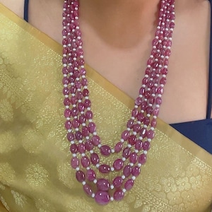 Puede incluir: Collar de varias hebras con cuentas de rubí ovaladas y pequeñas perlas blancas. El collar se muestra sobre una tela dorada y una blusa azul oscuro.