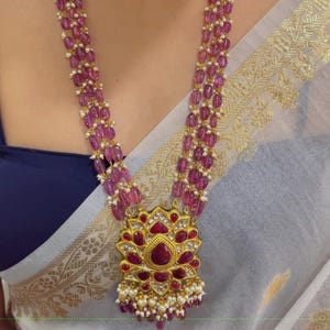 Puede incluir: Un collar de varias hebras con un colgante grande y pendientes a juego. El collar presenta cuentas rosas y un colgante dorado con piedras rojas y transparentes. Los pendientes tienen un diseño similar. La mujer lleva un top azul y una bufanda blanca.