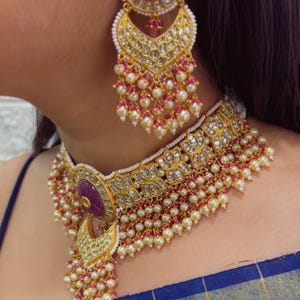 Puede incluir: Juego de collar y pendientes dorados con una piedra central de color rubí y detalles de perlas. La joyería presenta intrincados filigranas y un diseño en capas, con racimos de perlas colgantes.