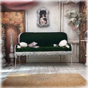Może przedstawiać: Miniaturowa sofa z ciemnozielonego aksamitu z białą metalową ramą i ozdobną koronką u podstawy. Sofa posiada dwie małe białe szydełkowane poduszki oraz dwie miniaturowe książki na siedzisku, a nad nią znajduje się portret w srebrnej ramie.