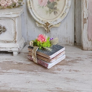 Könnte beinhalten: Ein Stapel aus drei quadratischen Miniaturbüchern, die mit einem goldglitzernden Band zusammengebunden sind. Das oberste Buch hat einen dunklen Einband und ist mit einem Bündel aus leuchtend rosa Kunstblumen und grünem Moos verziert.