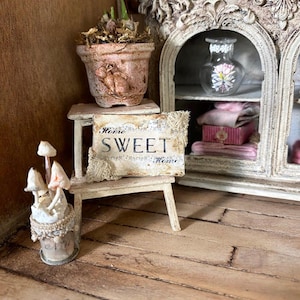 May include: A miniature scene featuring a small terracotta pot with a rabbit relief, a wooden stool holding a sign that reads Home SWEET Home, a glass vase with floral decals, and a small container with three white mushroom-shaped ornaments.