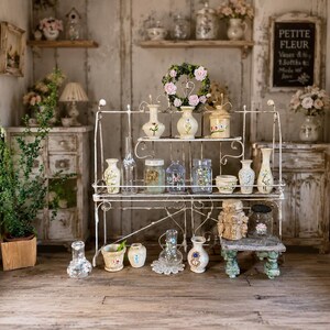 May include: A miniature room display featuring a white metal tiered shelf filled with various small ceramic vases, glass jars, and decorative items. A floral wreath sits on the top shelf, and a chalkboard sign reads PETITE FLEUR.