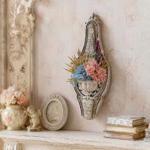 Könnte beinhalten: Eine dekorative Wandplatte mit einer zentralen Urnenform, gefüllt mit einem Arrangement aus künstlichen Blumen in Rosa-, Blau- und Violetttönen. Die Platte hat ein verziertes, verwittertes cremeweißes Finish mit detaillierten Reliefs.