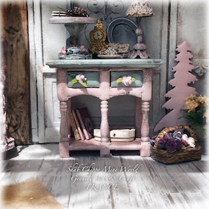 Könnte beinhalten: Ein Miniatur-Konsolentisch aus Holz in verwittertem Rosa und Hellblau mit zwei Schubladen und Blumenknöpfen. Auf dem Tisch stehen ein Tablett auf einem Fuß, eine goldene Uhr und eine Lampe. Darunter liegen Bücher und eine Schachtel. Text: Shab Chippy Mini World Genuine Art & Craft FRANCE.