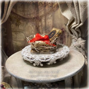 Könnte beinhalten: Ein Miniatur-Zweigkorb mit einem kleinen Buch mit dunklem Einband und goldener Schrift, das mit einem leuchtend roten Band zusammengebunden ist. Der Korb steht auf einer runden, weiß spitzenbesetzten Unterlage. Der Text "Shab Chippy Mini World Genuine Art & Craft FRANCE" ist sichtbar.