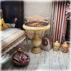 Könnte beinhalten: Ein runder Miniatur-Tisch mit einem abgenutzten cremefarbenen Finish und einer dunklen Tischplatte mit Blumenmuster. Der Tisch steht auf einem zentralen Säulenfuß. Der Text "Shab Chippy Mini World Genuine Art & Craft FRANCE" ist sichtbar.