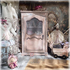 Könnte beinhalten: Ein Miniatur-Kleiderschrank aus Holz mit abgenutzter rosa Oberfläche, ausgestattet mit einem oberen Türpaneel aus Drahtgeflecht und einem kleinen Metallknopf. Das Stück wird neben einem Miniaturkleid auf einem Ständer, einer Spitzenpuppe und einem kleinen Wagen präsentiert.