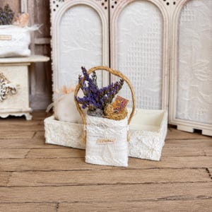 Könnte beinhalten: Ein Miniatur-Korb aus weißem Stoff mit einem Jute-Henkel, gefüllt mit getrockneten violetten Lavendelzweigen und einem kleinen rechteckigen Schild mit der Aufschrift JOURNAL. Der Korb ist mit weißer Spitze verziert und steht vor einem weißen Paravent.