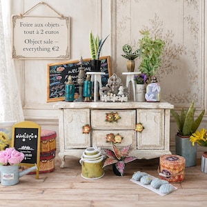 Könnte beinhalten: Ein weißer Miniatur-Holzschrank mit Blumenknöpfen, umgeben von verschiedenen kleinen Topfpflanzen, dekorativen Schildern mit Text, einem Sonnenblumenbild und kleinen Behältern. Ein Schild trägt die Aufschrift: Foire aux objets – tout à 2 euros.