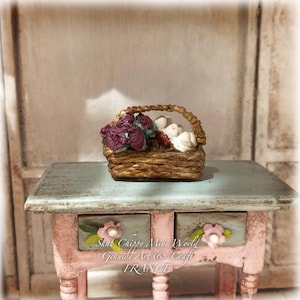 Könnte beinhalten: Ein kleiner, geflochtener brauner Korb mit violetten und weißen Blumen, der auf einem hellblau und rosa bemalten Holztisch steht, welcher zwei Schubladen mit jeweils einem rosa Blumenknopf und grünen Blattakzenten besitzt.