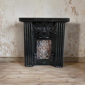 Può includere: Una cornice per camino in ghisa nera con colonne verticali scanalate e un fregio ornato da volute. Il pannello centrale presenta un rilievo di due figure che fiancheggiano un emblema circolare, con una griglia in filo metallico bianco alla base.