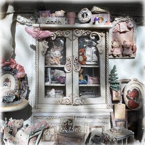 Może przedstawiać: Miniaturowa biała drewniana szafka z ozdobnymi rzeźbieniami i szklanymi drzwiczkami, prezentująca małe figurki zwierząt, pluszowego misia i pościel. Szafka jest otoczona różnymi miniaturowymi przedmiotami dekoracyjnymi, w tym oprawionymi portretami zwierząt.