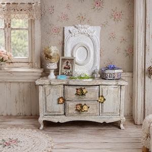 Könnte beinhalten: Ein Miniatur-Sideboard aus Holz mit abgenutztem weißem Finish, ausgestattet mit zwei zentralen Schubladen und zwei Seitenschränken. Die Schubladengriffe sind mit kleinen Blumenakzenten verziert. Auf dem Sideboard stehen ein weißer ovaler Rahmen, ein kleines gerahmtes Bild, eine dekorative Kugel auf einem Sockel und eine gemusterte Schachtel.