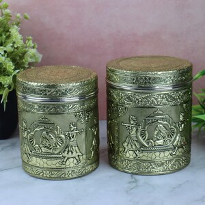 May include: Two antique gold-coloured metal canisters with intricate embossed designs. The cylindrical containers feature detailed scenes of figures and floral patterns. The lids and rims have a silver-coloured finish.