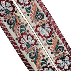 Peut inclure: Deux bandes de galon en tissu doré avec des broderies florales rouges, vertes et orange. Le galon présente un motif répétitif de fleurs avec des accents de paillettes argentées.
