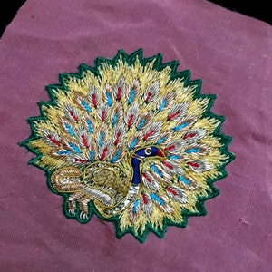 Könnte beinhalten: Ein detailliertes gesticktes Pfauenmotiv mit einem Farbschema aus Grün, Gold, Silber, Blau und Rot. Die Schwanzfedern des Pfaus sind fächerförmig ausgebreitet, und der Kopf des Vogels ist zur Seite gedreht.