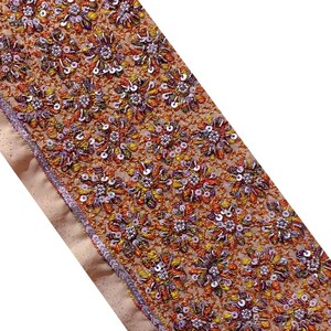 Può includere: Un bordino in tessuto beige con un motivo floreale ricamato con paillettes in tonalità arancio, giallo, viola e bianco.