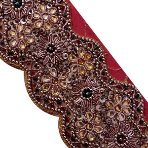 Op de afbeelding: Een close-up van een kralenbies met een gegolfde rand. De bies is gemaakt van gouden, bruine en roze kralen en pailletten. De bies is op een rode achtergrond.