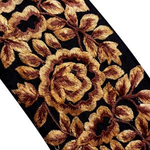 Könnte beinhalten: Nahaufnahme eines schwarzen Stoffes mit aufwendiger Stickerei. Das Design zeigt goldbraune Rosen und Blätter, die ein Blumenmuster bilden. Die Stickerei ist dicht und reichhaltig, mit leichtem Glanz.