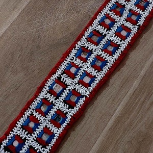 May include: A long, beaded belt with a red, white and blue geometric pattern. The beads are small and white, and the pattern is made up of squares and rectangles.
