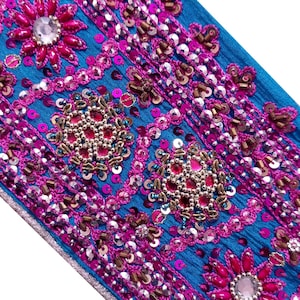 May include: Close-up of a vibrant blue textile adorned with intricate embroidery. The design features pink and magenta floral patterns, embellished with sequins and beads. The edges are finished with a silver trim, creating a luxurious and detailed appearance.