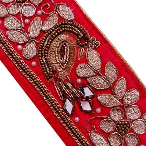 May include: Close-up of a red fabric strip with intricate embroidery. The design features gold and silver floral patterns, a peacock motif, and small mirrored accents. The edges are trimmed with gold thread and small white beads.