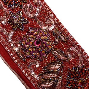以下が含まれることがあります： 赤色の生地のストリップのクローズアップ。スパンコール、ビーズ、刺繍された花の模様が施されています。デザインは、虹色と宝石色のアクセントを持つ星と花のモチーフが特徴です。エッジはサテンのボーダーで仕上げられています。