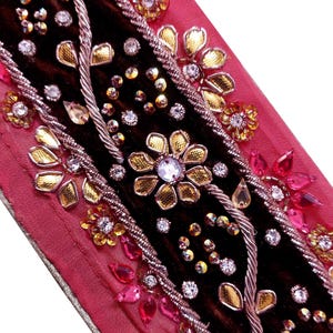 May include: Close-up of a decorative textile with a dark brown central band, bordered by pink fabric. The textile is embellished with gold and pink floral designs, clear and pink gemstones, and silver-coloured twisted rope.