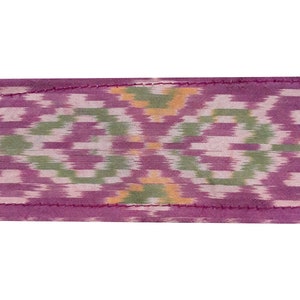 May include: A close-up of a purple fabric with a green and white ikat pattern. The fabric has a pink stitched edge.
