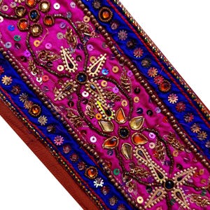 以下が含まれることがあります： 鮮やかなマゼンタ色の中央パネルに、複雑な金色の刺繍、多色のスパンコール、ファセットカットされた琥珀色のガラス石が施された装飾用ファブリックトリム。縁には金色のビーズのアクセントが付いた深い青色の帯があしらわれています。