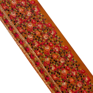 Könnte beinhalten: Ein diagonaler Stoffstreifen in einem warmen, goldbraunen Farbton. Der Stoff ist reich mit einem floralen Muster in Rot-, Orange- und Grüntönen bestickt. Ein dünner Goldrand verläuft entlang der Kante und verleiht dem Design einen Hauch von Eleganz.