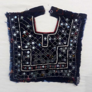 May include: A dark blue fabric neckpiece with a decorative design of small round mirrors and white embroidery. The fabric has a frayed edge and a red and white trim.