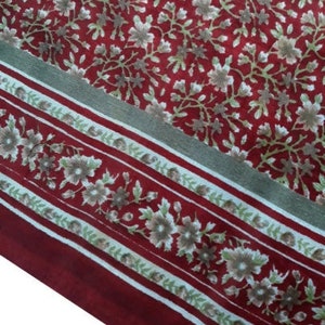 May include: Red fabric with a white floral pattern. The fabric has a border with a repeating pattern of white flowers and green leaves on a red background.