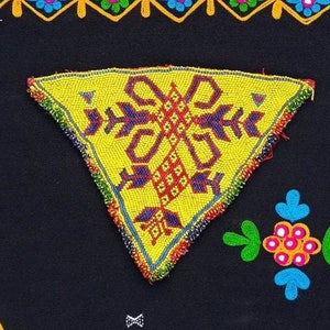 Puede incluir: Un parche triangular de tela amarilla con un diseño de cuentas de colores, cosido sobre un fondo de tela negro. El parche presenta un patrón geométrico con cuentas rojas, azules y amarillas.