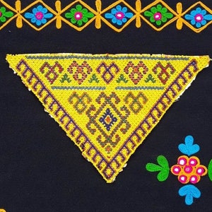 Puede incluir: Una tela negra con diseños bordados de colores. Un triángulo amarillo con un patrón geométrico se encuentra en el centro. La tela tiene un borde a rayas.